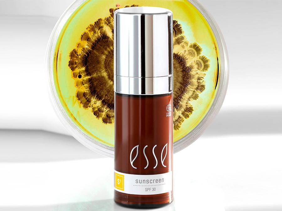 A brown bottle with a silver pump labeled "sunscreen SPF 30" stands in front of a round petri dish showcasing a colorful, intricate growth pattern. The bottle has the brand name "esse" in white text. The background is white, emphasizing the product and dish.