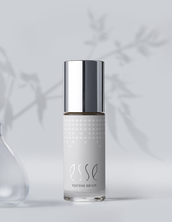The minimalist Esse Ageless Serum bottle with a silver cap rests on a light grey surface, casting soft shadows against a blurred floral pattern in the background.