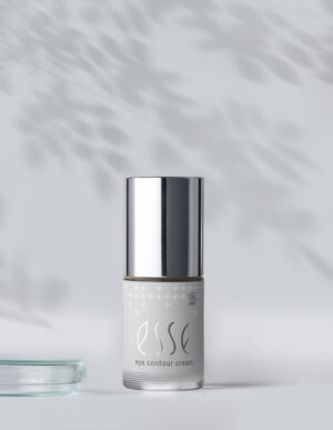 A small bottle of Esse Eye Contour Cream with a shiny silver cap stands upright on a light surface, soft leafy shadows on the background. To the left, a clear glass dish is partially visible.