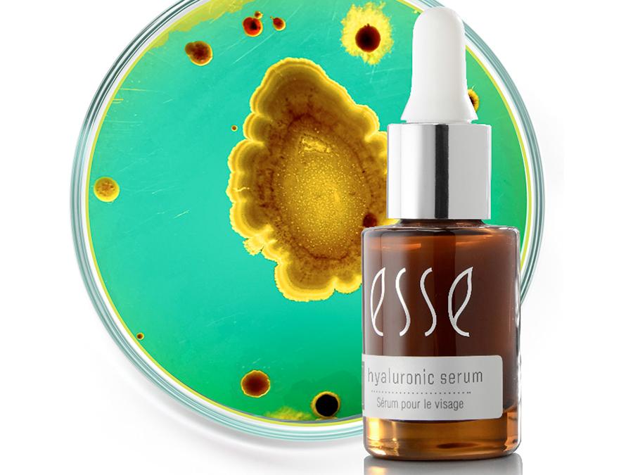 A small brown glass bottle labeled "Esse Hyaluronic Serum" with a white dropper cap is positioned in front of a green and yellow petri dish with various circular patterns, suggesting a scientific or skincare-related context.
