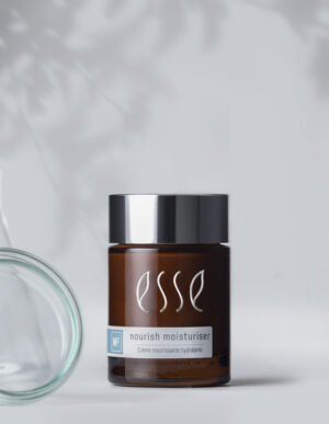 A dark brown glass jar with a black lid, bearing the labels "Esse Nourish Moisturiser" and "Crème nourrissante hydratante," rests on a light gray surface adorned with faint leaf shadows in the background.