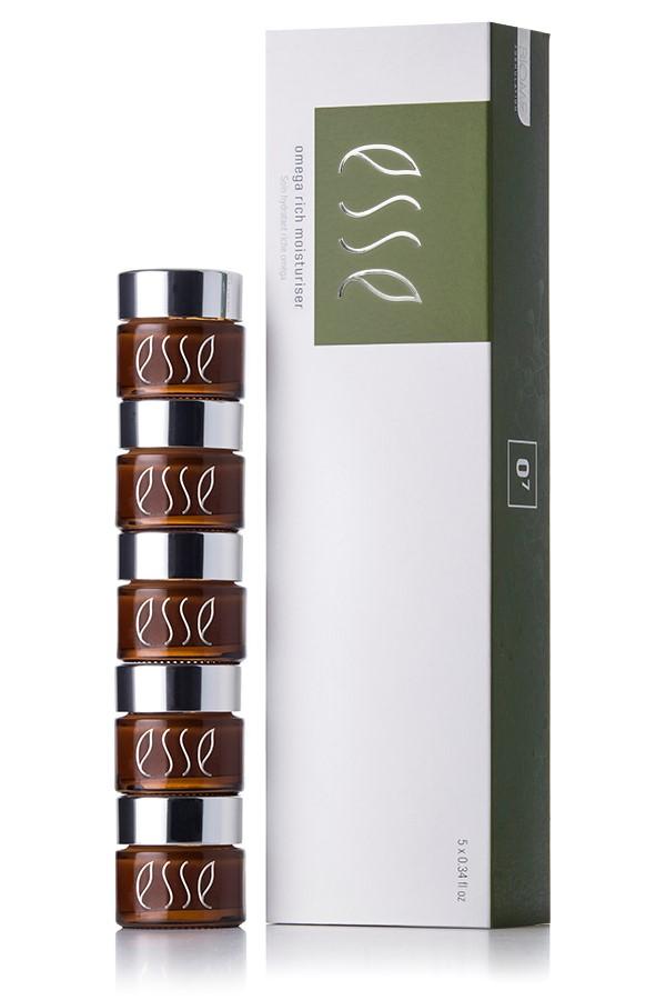 A stack of four brown "Esse Omega Rich Moisturiser" cosmetic jars with silver lids are placed beside a sleek white and green box featuring the same branding. The box stands upright on a white surface, showcasing the product packaging.