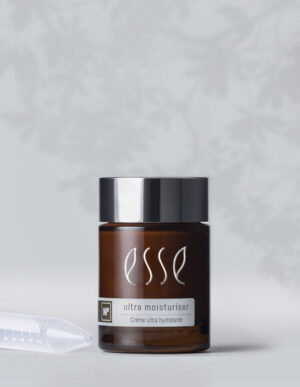 A brown jar of Esse Ultra Moisturiser with a silver lid, labeled "Creme ultra hydratante" and showcasing an "MB" logo. The jar is set against a light grey background accented by a soft, blurred floral pattern, with a clear plastic dropper lying beside it.