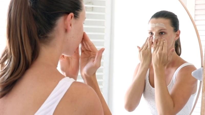 A woman with her hair in a ponytail is looking into a mirror, applying the Esse Microderm Exfoliator to her face. She is wearing a white tank top and is standing in a bright room with a round mirror and white louvered shutters in the background.