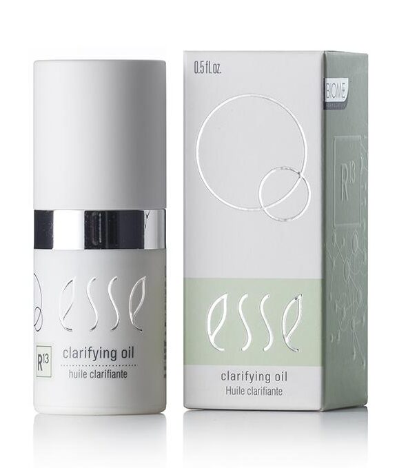 The image features a bottle of Esse Clarifying Oil along with its packaging. The cylindrical bottle is adorned with a white cap and a metallic silver band. The light green and white box showcases the product name and various branding elements. The bottle has a capacity of 0.5 fl oz.