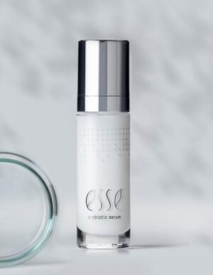 A sleek bottle of Esse probiotic serum with a metallic pump dispenser stands upright. The branding text "Esse probiotic serum" is visible on the frosted glass body. The cap and background are shades of gray and white, giving the image a clean, minimalistic look.