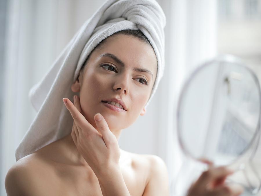 A person with a towel wrapped around their head gazes into a handheld mirror, gently touching their face. The background is softly lit, giving a serene, self-care ambiance.