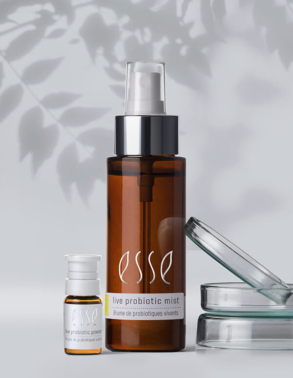 Two brown Esse Live Probiotic Mist bottles sit on a white surface near a clear glass lid and dishes, with soft leaf shadows behind them. The larger bottle features a pump dispenser, while the smaller one has a white cap.