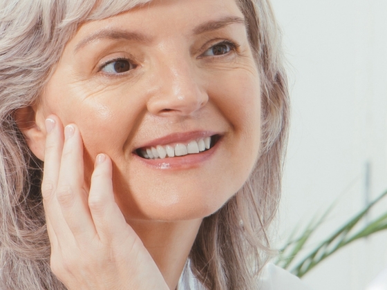 A smiling older woman with light skin and gray hair touches her cheek gently, appearing content. A green plant is visible in the blurred background.