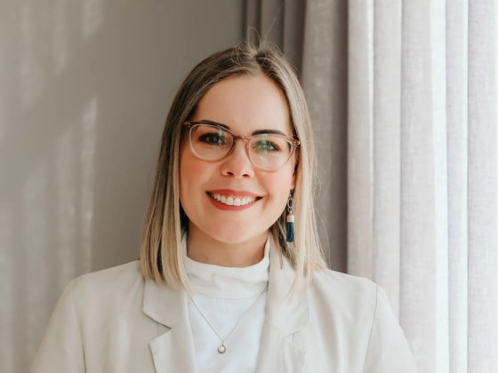 A woman with straight, shoulder-length blonde hair and glasses smiles in a white blazer and top, accessorized with a necklace and dangling earring, standing before light curtains—ready to celebrate the Legacy Probiotic Skincare.