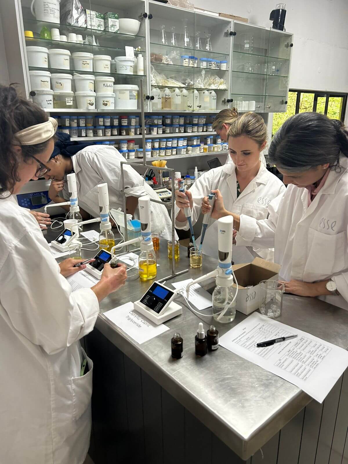 Five people in white lab coats work at a laboratory counter with scientific instruments and bottles. Shelves behind them hold jars and containers of chemicals. The workspace is organized and well-lit.