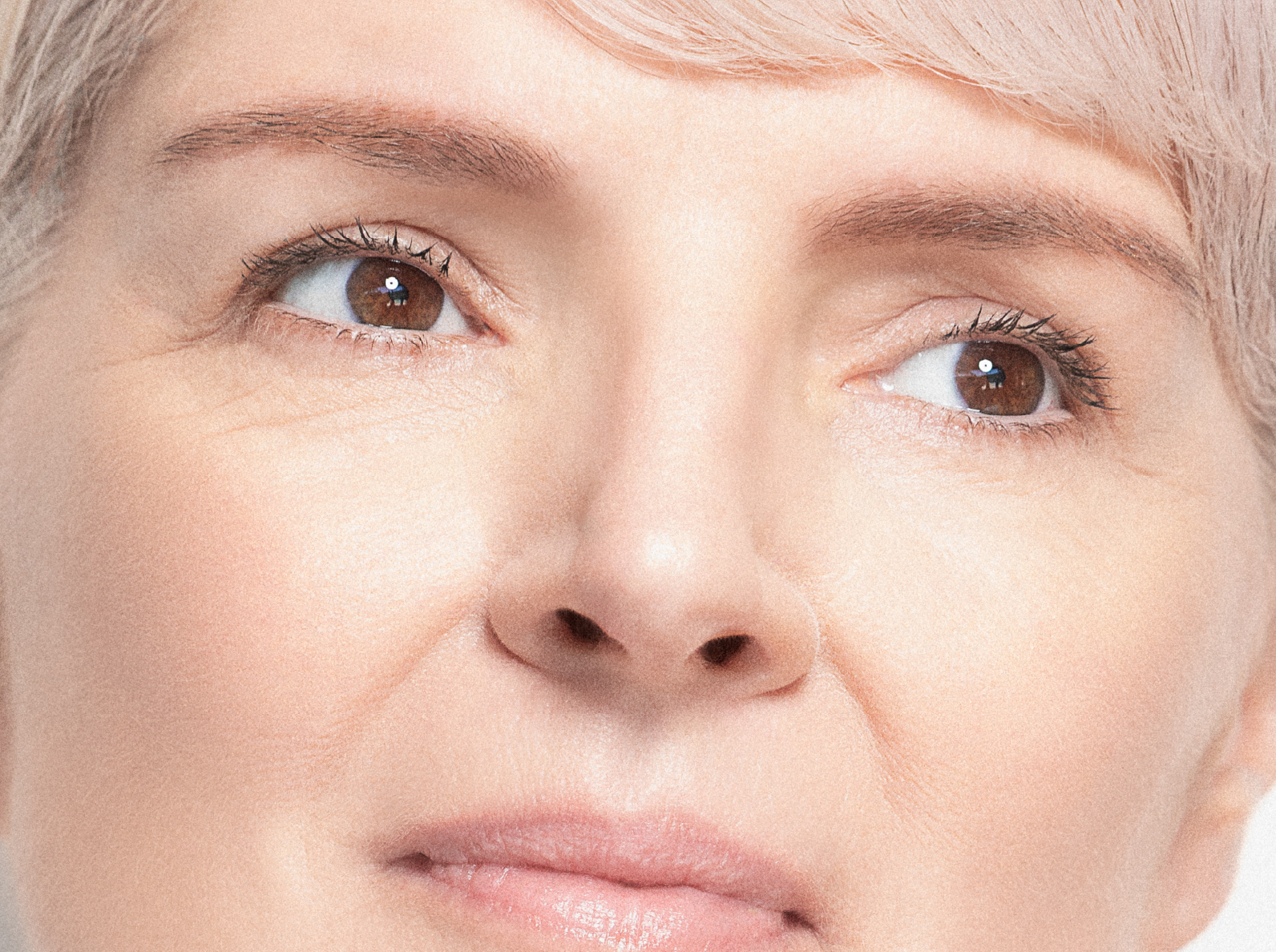 Close-up of a middle-aged person with light skin and short, light-blonde hair, showing smooth skin, brown eyes, and subtle facial lines, looking off to the side with a calm expression.