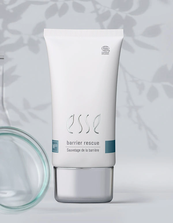 A white tube of Esse Barrier Rescue stands upright on a light surface, accompanied by the Esse Bakuchiol Serum, with a blurred glass object and leaf shadows in the background.